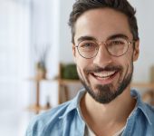 A smiling man with glasses and a beard wears a denim shirt. He stands in front of blurred shelves.
