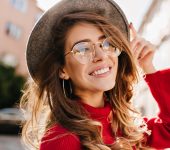 Woman in a red sweater smiles while touching her hat. She is wearing round glasses and hoop earrings, standing outdoors with buildings in the background.