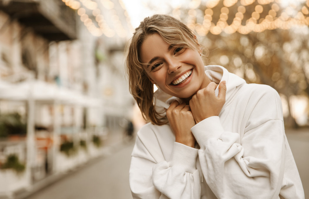 Person in a white hoodie smiling outdoors, with blurred lights and buildings in the background.