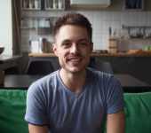 A man with short dark hair and a trimmed beard is smiling while sitting on a green couch in a modern kitchen.