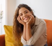 A woman with long brown hair, wearing a beige shirt, sits on an orange sofa and smiles at the camera with her hands resting on her face.
