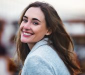 A woman with long brown hair and a light gray coat smiles at the camera outdoors, with a blurred background.