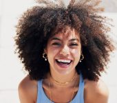 A person with curly hair, wearing a blue tank top and layered necklaces, smiles outdoors in bright sunlight.