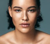 A young woman with curly hair and freckles looks directly at the camera against a plain, neutral background. She has a calm, neutral expression and bare shoulders.