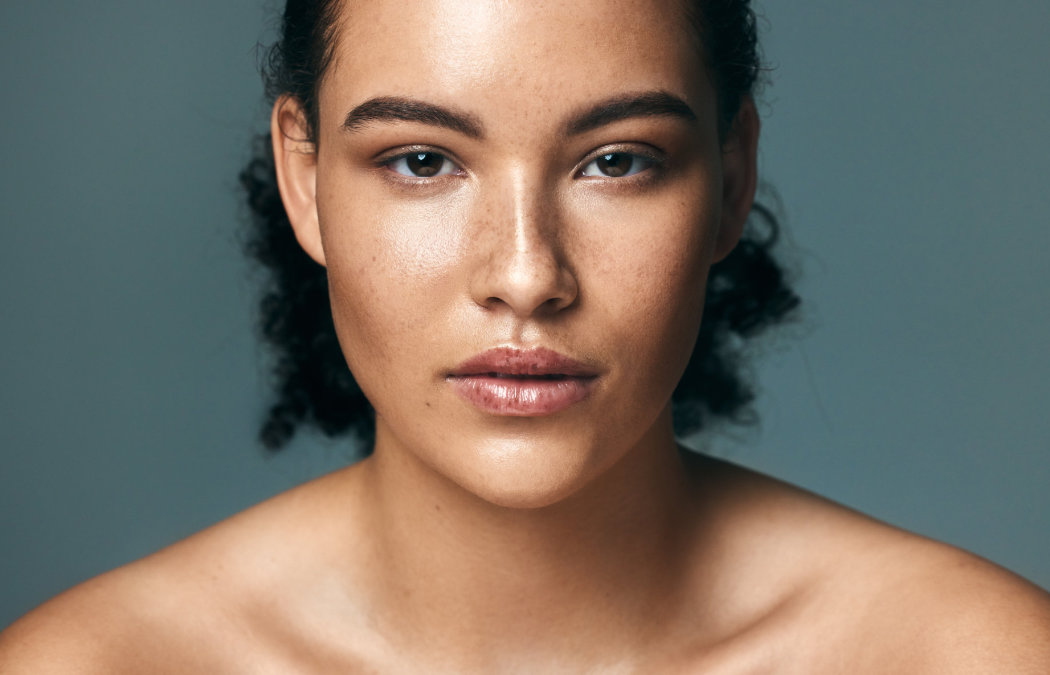 A young woman with curly hair and freckles looks directly at the camera against a plain, neutral background. She has a calm, neutral expression and bare shoulders.