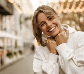 Person in a white hoodie smiling outdoors, with blurred lights and buildings in the background.