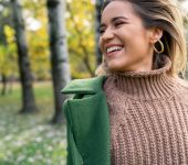 A person wearing a brown knit sweater and green coat stands outdoors, smiling, with blurred trees and grass in the background.