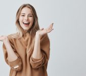 Pretty beautiful woman with blonde long hair looking at camera having excited and happy facial expression, clapping with her hands against blank studio wall, expressing her excitement with present