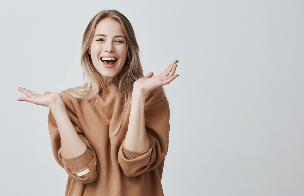 Pretty beautiful woman with blonde long hair looking at camera having excited and happy facial expression, clapping with her hands against blank studio wall, expressing her excitement with present