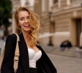 Beautiful smiling woman in elegant black suit posing outdoor in old european city. Blond wavy hairs, perfet skin.